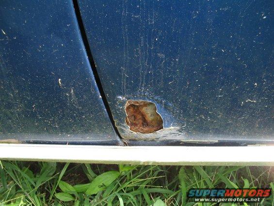 Close-up of rust hole on the bottom edge of a blue car door, with grass below