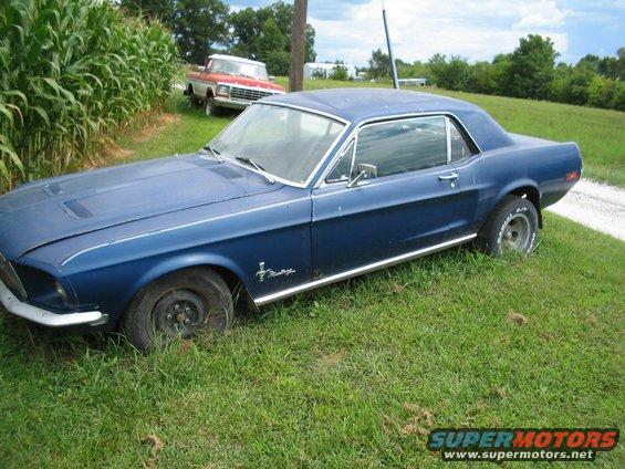 Blue 1967 Ford Mustang coupe parked in grass, with a red and white pickup truck in the background