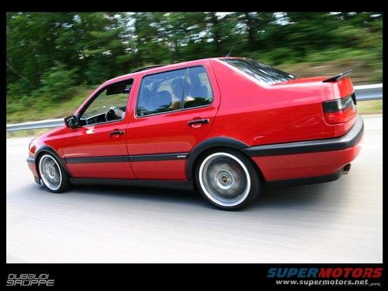 Red sedan with aftermarket wheels driving on a road, motion blur visible