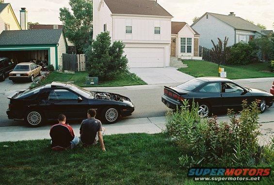 1989 Mazda RX-7 with hood open and a second black coupe parked on street, two people sitting on grass