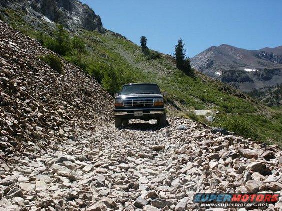 1996 Ford Bronco driving on a steep, rocky mountain road with sparse trees and a clear blue sky