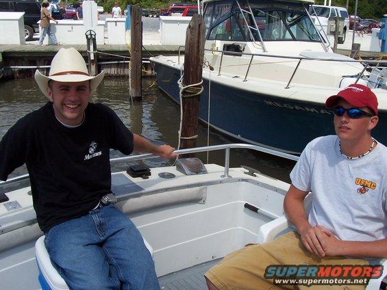 Two men on a boat, one wearing a cowboy hat, with a larger boat behind them