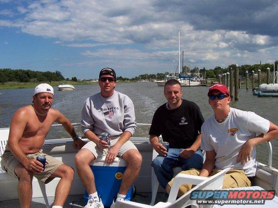 Four guys on a boat, some holding cans, with boats docked in the background