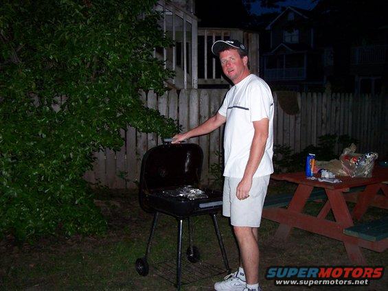 guy in white shirt and shorts by a black charcoal grill in a backyard at dusk