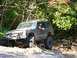 1990 Ford Bronco - A dark-colored Jeep Cherokee XJ navigates a challenging off-road course through a wooded area.