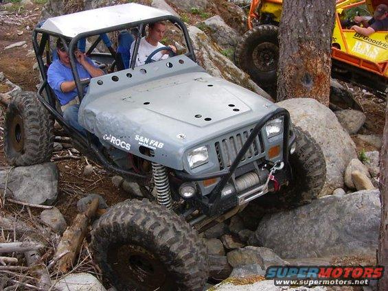 Gray custom off-road vehicle with roll cage navigating rocks. Text on side reads 'ROCS S&N FAB'