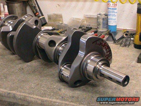 Engine crankshaft on a workbench with tools and cans in the background. Some text is visible on the crankshaft