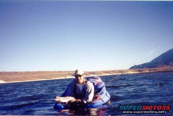 Man in blue raft holding a fish, with a mountain and clear blue sky in the background