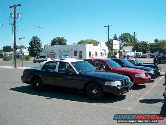 Several police cars parked in a lot, including a black and white unit with American flags on top