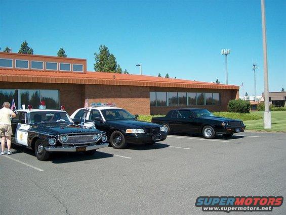 Three police cars parked in a lot: a vintage black and white sedan, a black Ford Crown Victoria, and a black coupe