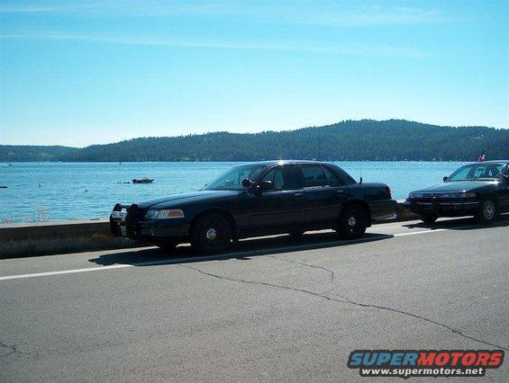 Two black Ford Crown Victorias parked on a roadside next to a lake