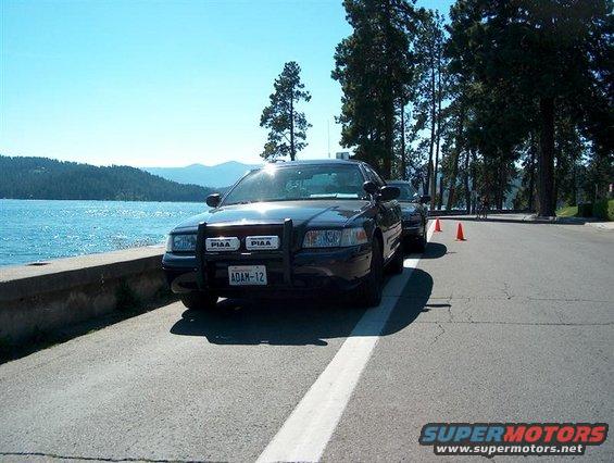 Black police car with PIAA lights and "ADAM-12" plate parked by a lake