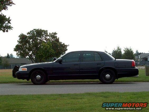 Dark blue 2001 Ford Crown Victoria police car, side view, parked on a street with a tree and houses in the background