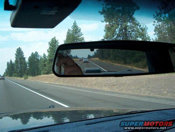 View from inside a car, looking at the rearview mirror which reflects a highway with other cars and trees. A hand is visible in the mirror