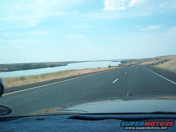Driving down a highway next to a lake under a blue sky with clouds. Part of a dashboard is visible in the foreground