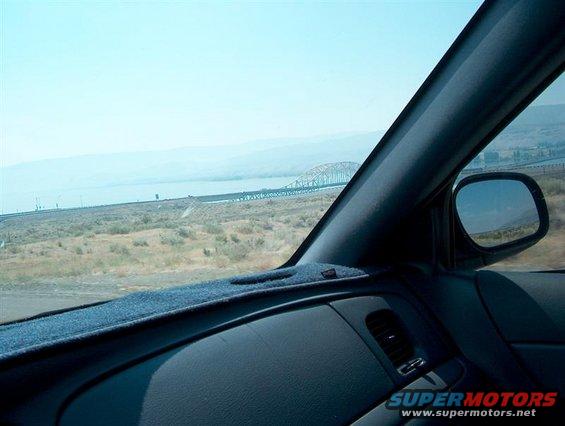 Car interior dashboard and side mirror showing a bridge and dry landscape under a clear sky
