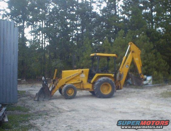 Yellow John Deere backhoe loader parked on dirt, with trees and a metal building in the background
