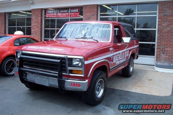 Red 1981 Ford Bronco with 'MAGNOLIA ROAD FIRE COMPANY' text on the side, parked in front of a building. A red sports car is visible to the left