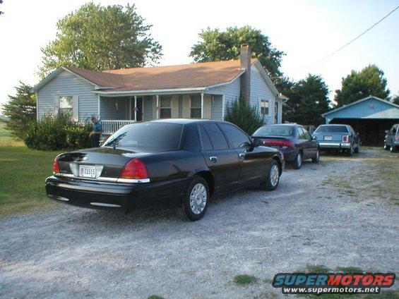 Black sedan parked in front of a house, with other cars behind it. License plate is visible