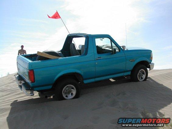 Teal Ford Bronco with a red flag on a sand dune, driver's side visible
