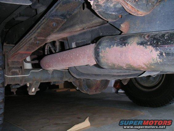 Underside view of a vehicle's rear axle, leaf springs, and exhaust system. Shows rust and wear on components