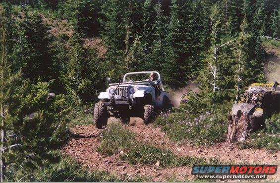 White Jeep CJ-7 driving on dirt trail in wooded area, kicking up dust