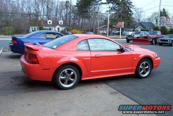 Orange 2004 Ford Mustang parked next to a blue car, with other vehicles and buildings in the background