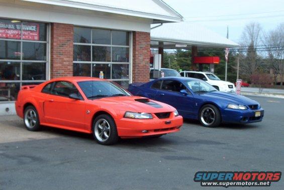 Orange 2004 Ford Mustang parked in front of a brick building, with a blue Mustang behind it