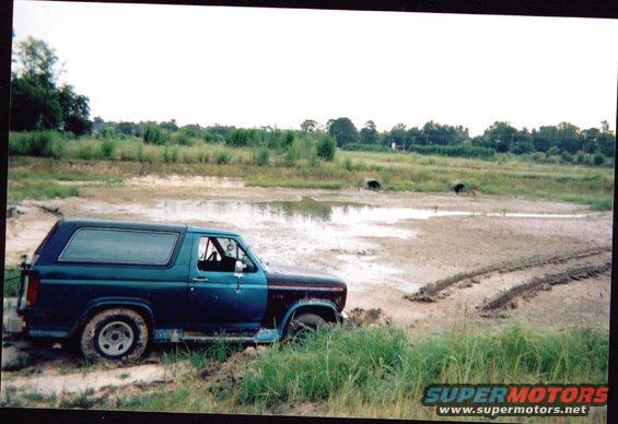 Side view of a 1985 Ford Bronco stuck in a muddy field with tire tracks visible