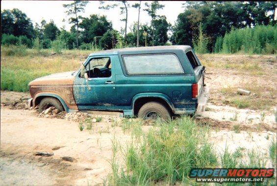 Green and rusty Ford Bronco stuck in mud, front driver's side wheel submerged