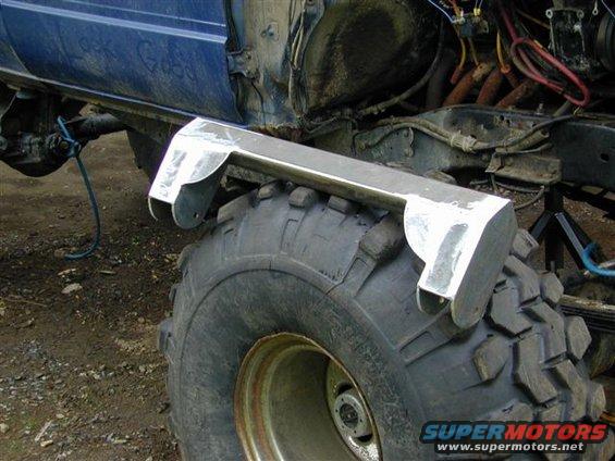 Side view of a heavily modified truck chassis with large off-road tires and custom metalwork near the wheel well. Some writing is visible on the blue body panel