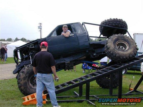Black modified truck with large tires on a ramp, driver in cab, man in red hat watching