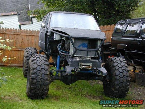 Custom off-road vehicle with large tires, exposed engine, and roll cage. Another black SUV is parked next to it