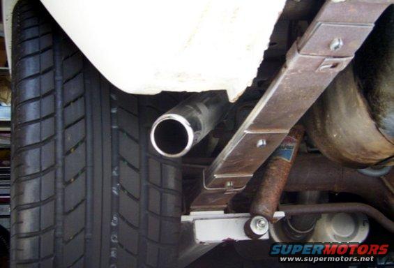 Close-up of a car's rear leaf spring, shock absorber, tire, and exhaust pipe. The shock absorber has a blue and red label