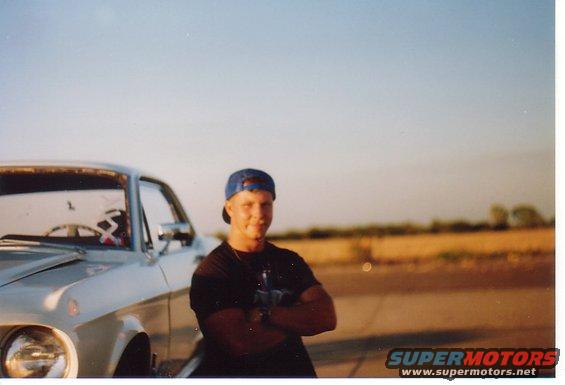 Guy in a blue baseball cap and black t-shirt leans against a silver classic car. Field and sky in background