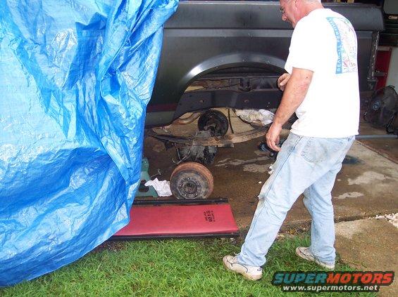 Dark green Ford Bronco with rear axle removed, partially covered by a blue tarp. A man stands nearby