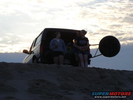 Van parked on sand dunes with people standing near the open rear hatch at sunset