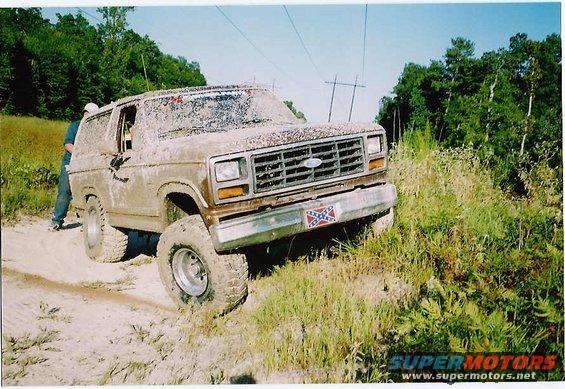 Mud-covered 1980s Ford Bronco with large tires on a dirt path, trees in background