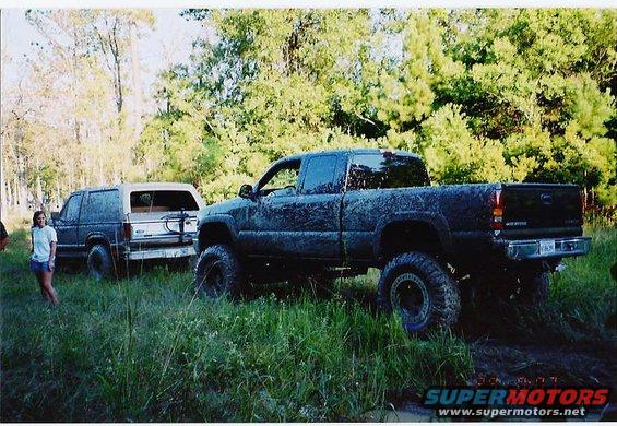 Muddy pickup truck and SUV parked in tall grass with trees in background