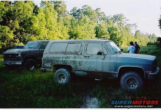 Two dirty SUVs parked in tall grass, one in foreground is blue-grey, the other behind it is dark brown