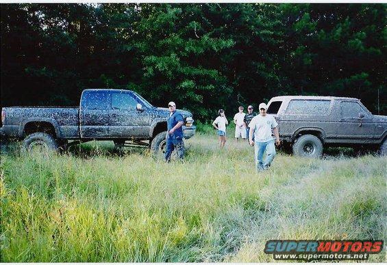 Mud-covered 1980s Ford Bronco and pickup truck parked in tall grass with several people standing around