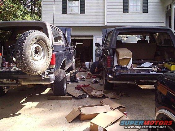 Two Ford Broncos parked in a driveway with hoods and tailgates open, one being worked on by a person
