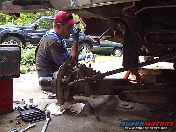 Man in red cap and overalls working on vehicle axle and suspension parts. Tools scattered on ground. Another SUV visible in background