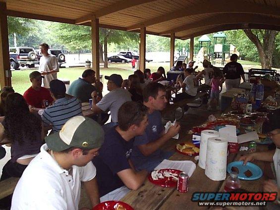 People eating at picnic tables under a covered pavilion, with a playground in the background