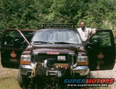 Maroon 2000 Ford Excursion with front winch and roof rack, driver's side door open, man standing beside it
