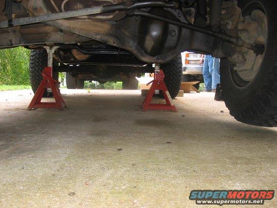 Vehicle underbody on jack stands, showing suspension and drivetrain components. Another vehicle is partially visible in the background