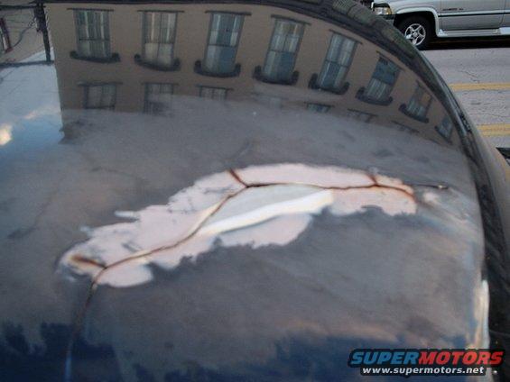 Cracked paint and primer on dark car roof; building reflection visible