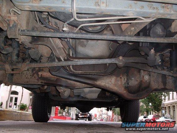 Underside of a vehicle with visible suspension and drivetrain components. A dark SUV is visible in the distance