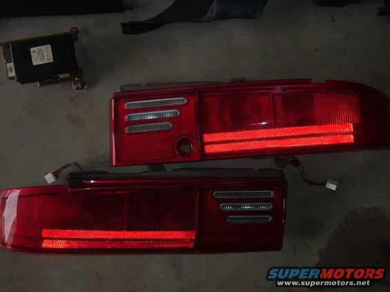 Two red car taillights, one above the other, with wiring harnesses attached. A black electronic module is visible to the left