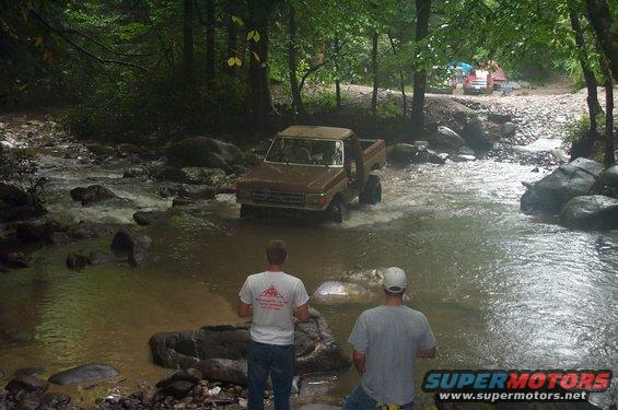 Brown pickup truck driving through a rocky stream, two men watching from the bank